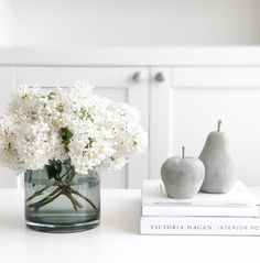 white flowers and pears in a glass vase on a table with two books next to it