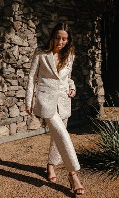 a woman standing in front of a stone wall wearing a white jacket and pants with high heels