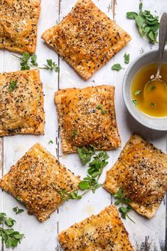 square crackers with herbs and olive oil on a white surface next to a bowl of dipping sauce