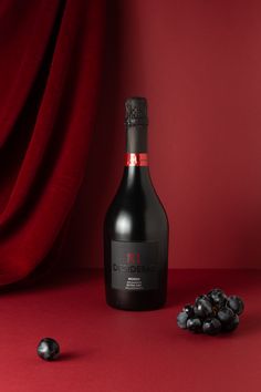 a bottle of red wine next to some black grapes on a red surface with a red curtain behind it