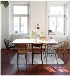 a dining room table with four chairs and a rug in front of two windows on the wall