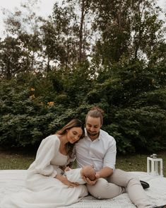 a man and woman sitting on top of a white blanket next to each other with trees in the background