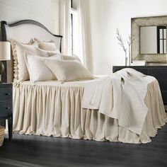 a white bed with ruffled bedspread in a bedroom next to a dresser and mirror