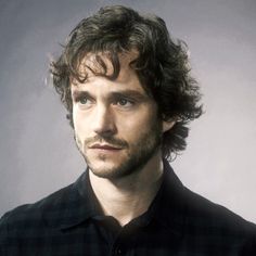 a man with curly hair wearing a black shirt and looking at the camera while standing in front of a gray background