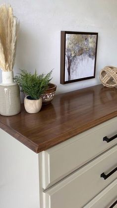 a white dresser topped with lots of drawers next to a painting and potted plant