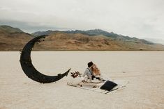 a woman sitting on top of a blanket next to a crescent shaped object in the desert