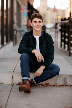 a young man is sitting on the sidewalk smiling at the camera while wearing jeans and a flannel shirt