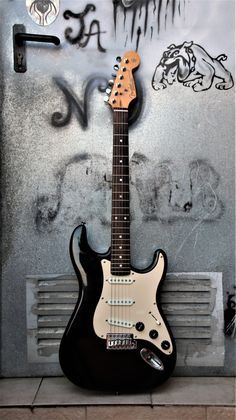 a black and white guitar sitting in front of a wall with some graffiti on it