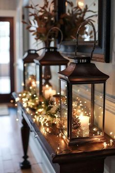 three lanterns are lit up on a mantle with christmas lights and greenery in front of them