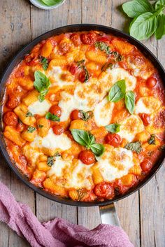 a skillet filled with pasta and sauce on top of a wooden table next to basil leaves