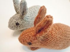 two knitted bunnies sitting next to each other on a white surface, one is brown and the other is gray