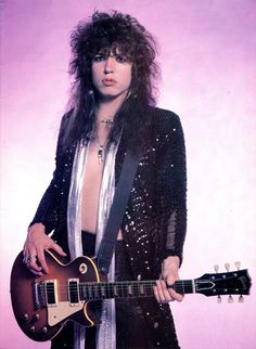 a man with long hair holding a guitar in front of a purple background and posing for the camera