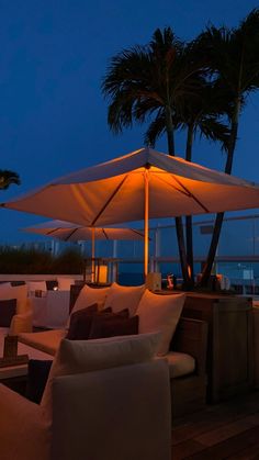 an outdoor seating area with umbrellas and palm trees at night, overlooking the ocean