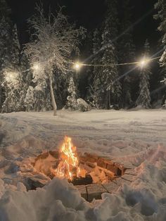 an open fire pit in the middle of snow covered ground with string lights above it