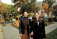 two women standing next to each other in front of pumpkins on the ground and trees