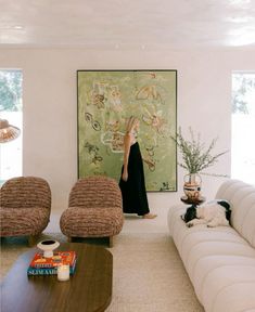 a woman standing next to a couch in a living room with two chairs and a painting on the wall