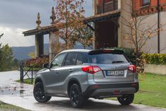 the rear end of a silver car parked in front of a building with trees and flowers