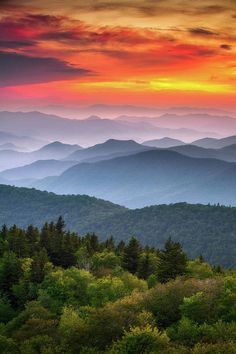 the sun is setting over mountains and trees in the foreground, as seen from an overlook