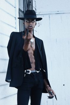 a man in a black suit and hat standing next to a white wall with his hand on his mouth