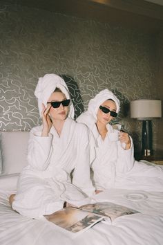 two women in bathrobes sitting on a bed and one is reading a magazine