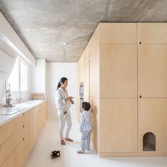 a woman standing next to a little boy in a room with wooden cabinets and drawers