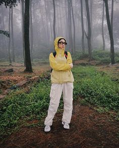 a woman standing in the middle of a forest wearing a yellow jacket and white pants