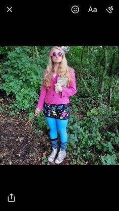 a woman in pink jacket and blue tights standing next to green bushes with trees