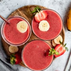 three glasses filled with smoothie and bananas on top of a wooden cutting board next to sliced strawberries