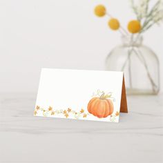 a white card with an orange pumpkin on it next to a vase filled with flowers