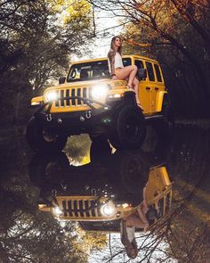 a woman sitting on top of a yellow jeep in the woods with trees reflected in the water