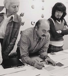 three people standing around a table with papers and pens in front of them, looking at something on the paper