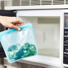 a person is holding a plastic container with broccoli in it near an open microwave door