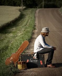 a man sitting on the side of a road next to a suitcase and skateboard