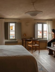 a woman sitting on a bed in a bedroom next to a desk and window with curtains