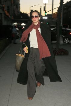 a woman is walking down the sidewalk carrying a purse and wearing a long black coat