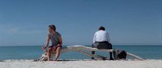 two people sitting on a bench made out of driftwood at the beach with water in the background