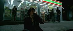 a woman sitting on the sidewalk in front of a store at night with people walking by