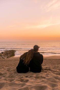 two people sitting on the sand looking out at the ocean and sunset in the distance