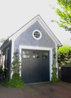 a gray garage with white trim and round window on the top floor is surrounded by greenery