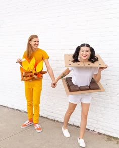 two women holding hands and standing in front of a white brick wall with an open cardboard box on their head