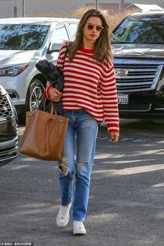 a woman walking down the street carrying a brown bag and wearing ripped jeans, a red striped sweater and white tennis shoes