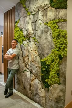 a man standing next to a wall covered in green mossy plants and rocks with his arms crossed