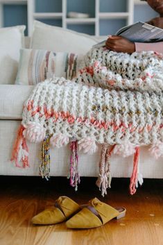 a woman sitting on a couch reading a book next to slippers and a crocheted blanket