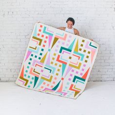 a man is holding up a large quilt in front of a white brick wall with an abstract pattern on it