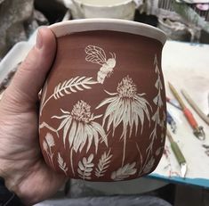 a hand holding a brown and white cup with flowers painted on the outside, in front of paintbrushes