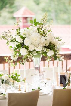 a table with white flowers and greenery in a tall vase on top of it