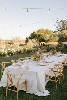 a long table with white linens and wooden chairs is set up for an outdoor dinner