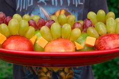 a man holding a tray with apples, grapes and oranges on it in front of him