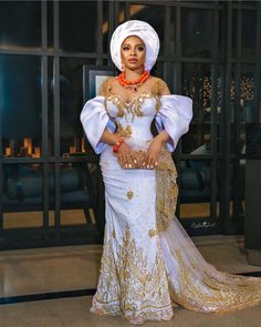 an african woman wearing a white and gold dress with orange accessories on her head, standing in front of a glass door