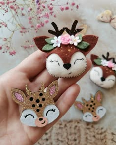 the hand is holding three little deer brooches with flowers on their head and one has eyes closed